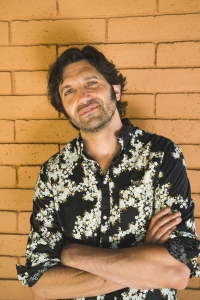 Man with dark hair and a beard stands against an orange brick wall, wearing a black shirt with a white floral pattern and crossing his arms.