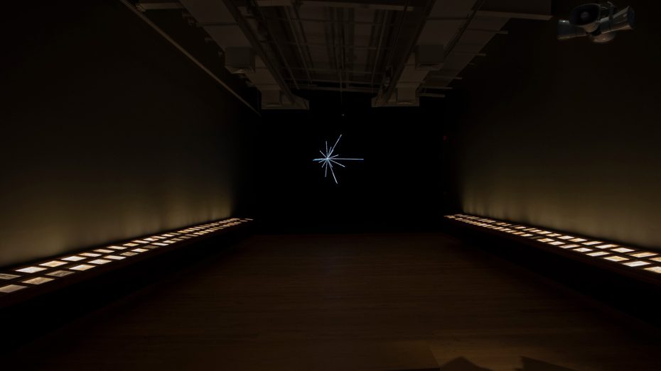 A dimly lit gallery room in museums, featuring illuminated display cases along both walls and a starburst light sculpture glowing at the far end.