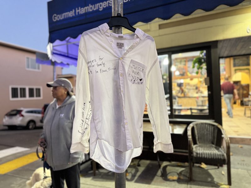 A white button-down shirt with handwritten messages, including “Justice for Bandy,” hangs on a pole on a sidewalk near shops and pedestrians.