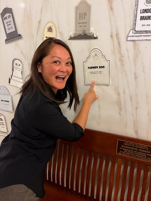 A woman smiles and points at a tombstone-shaped plaque on a wall labeled "Carmen Doo." Several other memorial plaques are also visible.