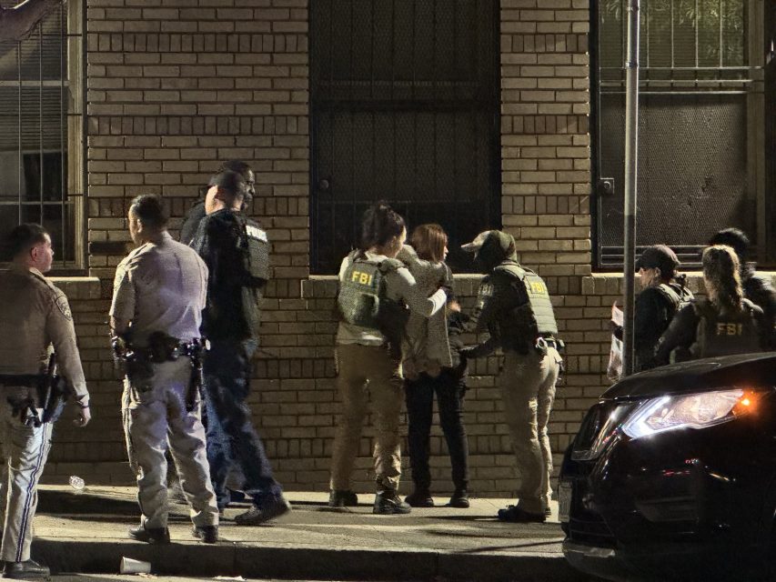 Law enforcement officers, including FBI agents, apprehend and search individuals on a sidewalk at night near a brick building.
