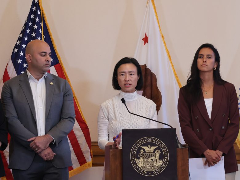 Three people stand at a podium with microphones, flanked by American and Californian flags, during a press conference.