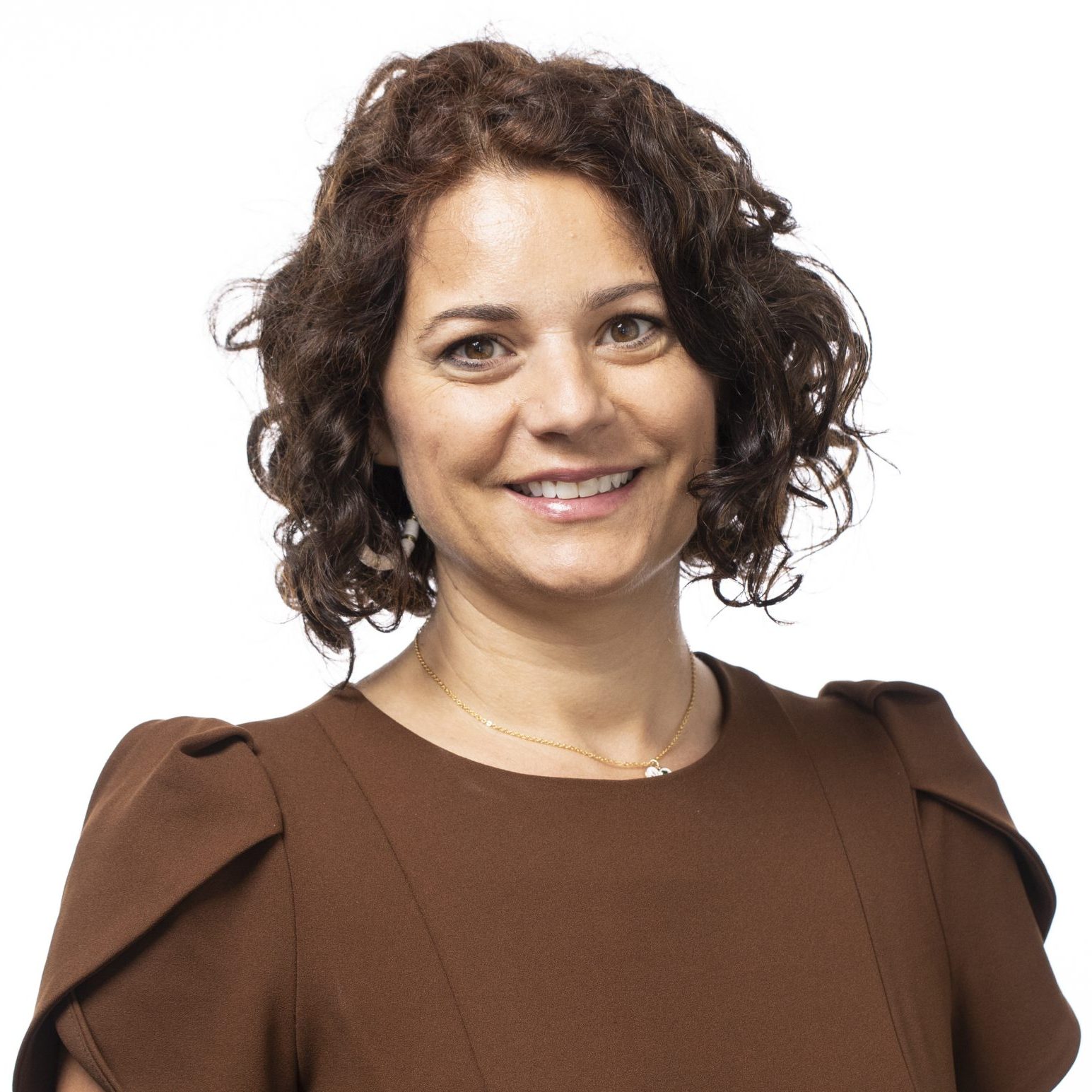 A woman with curly brown hair, wearing a brown dress, smiles at the camera against a plain white background.