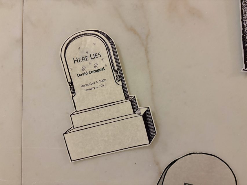 A paper cutout of a tombstone with text that reads: "Here Lies David Compost, December 4, 2008 - January 6, 2017," placed on a light-colored surface.
