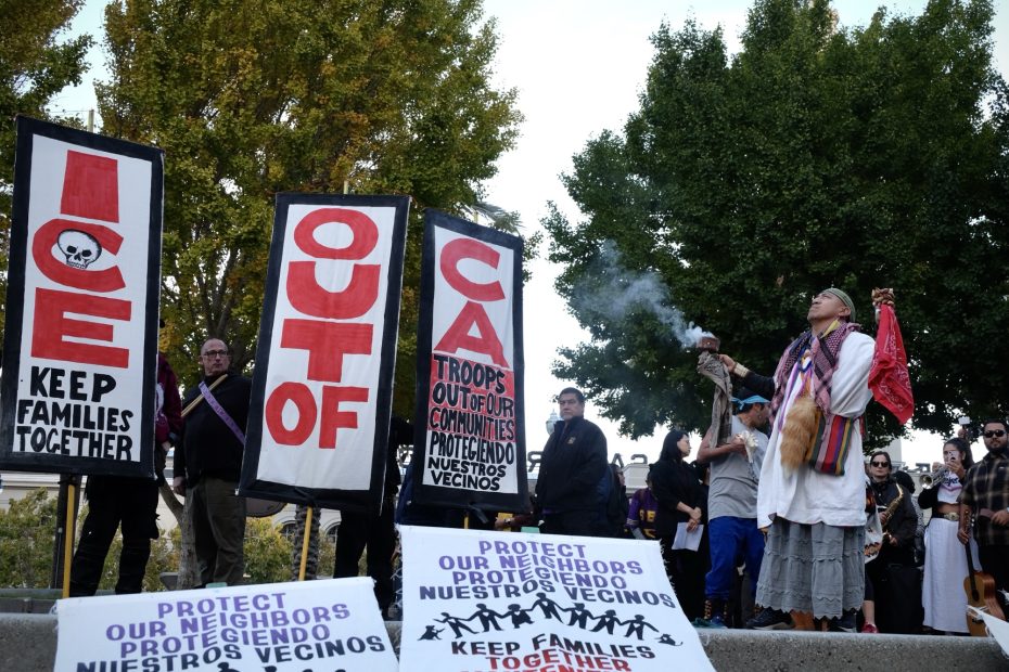 A group of people hold signs reading "ICE OUT OF CA" and "Keep families together" at a protest; a person on the right burns sage.