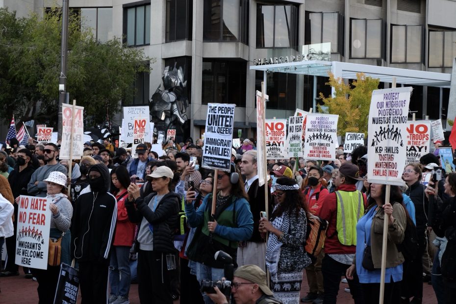 A large group of people gathers at a protest, holding signs with messages supporting immigrant rights, anti-racism, and unity, in an urban outdoor setting.