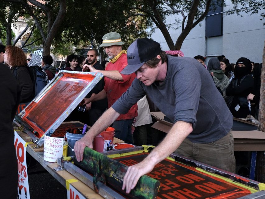 Two people stand at outdoor tables screen-printing protest signs, while others gather around. The signs read "No H8" and "NO HATE." Various art supplies are visible.