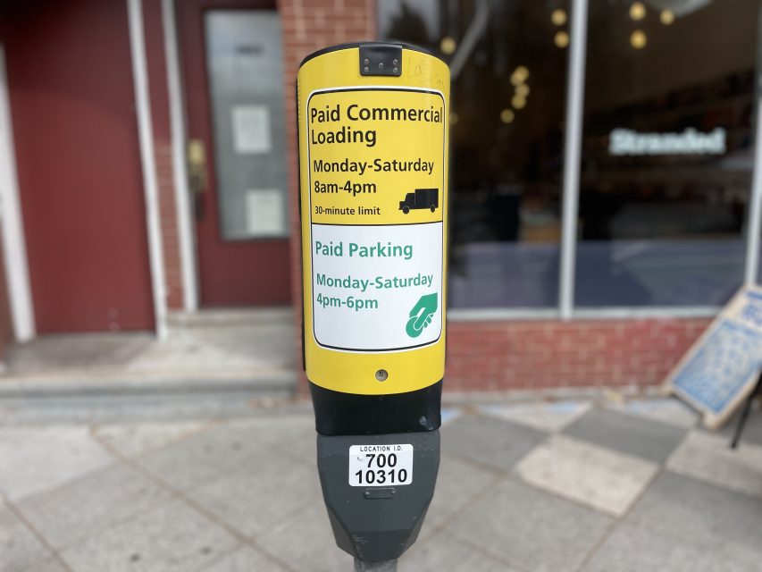 A parking meter with signs indicating paid commercial loading from 8am-4pm and paid parking from 4pm-6pm, Monday to Saturday, on a city sidewalk.