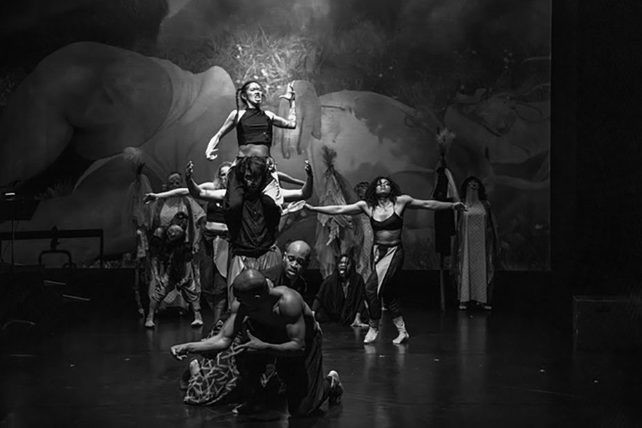 A group of dancers performs on stage in dramatic poses; one woman is lifted above others. The background shows a large mural of a reclining figure. The scene is in black and white.