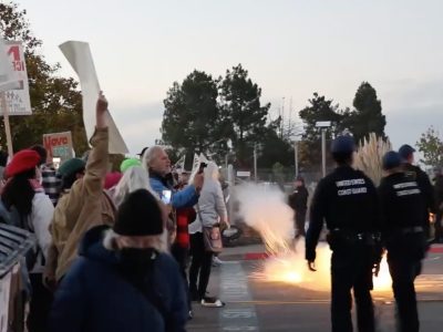 Protesters holding signs about immigration face police officers as a bright flare or pyrotechnic device emits sparks on the street between them.