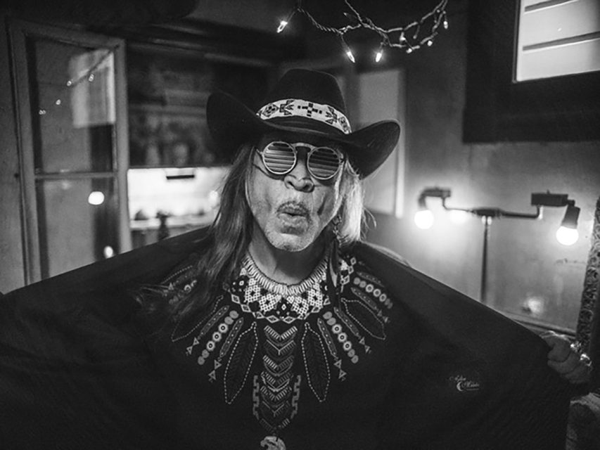 A person wearing a cowboy hat, patterned shirt, and striped sunglasses poses indoors with their arms out, in black and white lighting.