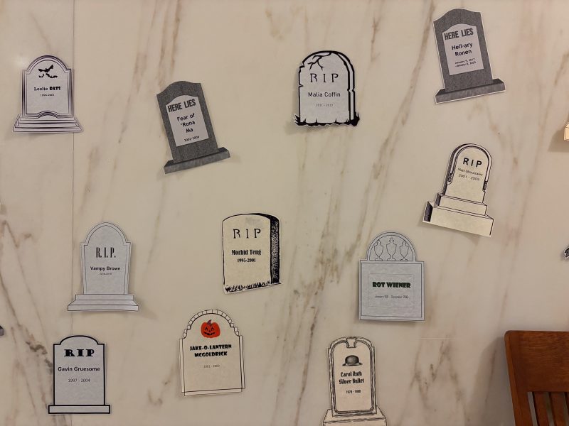 Paper cutouts of gravestones with humorous and fictional names and epitaphs are displayed on a beige marble wall.