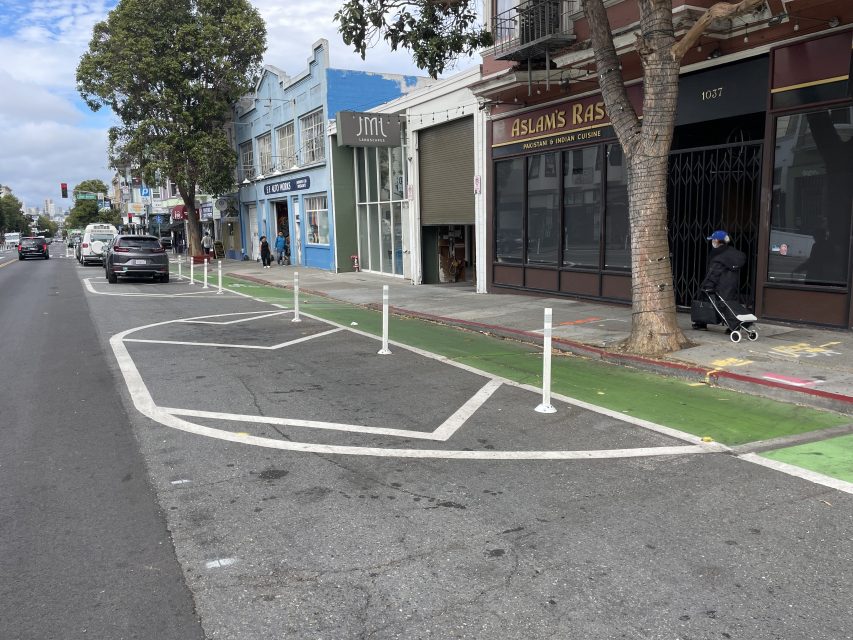 A bike lane protected by white posts runs along a city street with parked cars, shops, and a person with a stroller on the sidewalk.