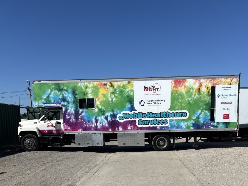 A large mobile healthcare services truck with a colorful tie-dye design and clinic logos is parked on a gravel lot under a clear blue sky.