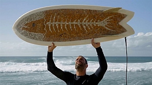 A man stands on the beach holding a surfboard above his head; the surfboard features a large fish skeleton design.