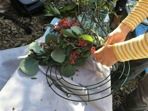 A person in a yellow-striped shirt arranges green leaves and red berries on a metal wreath frame atop a cloth-covered surface.