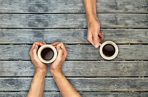 Two people holding cups of coffee sit across from each other at a wooden table, seen from above.