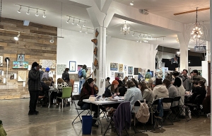 A group of people sit at tables and work on art projects in a bright, spacious gallery filled with artwork on the walls.