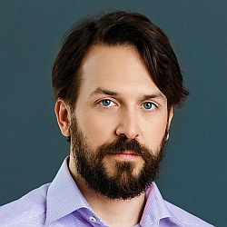 A man with medium-length brown hair, a full beard, and blue eyes is wearing a light purple collared shirt, posed against a plain dark background.