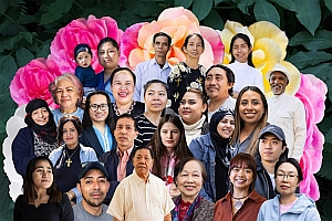 A group portrait of diverse people of various ages standing together in front of a background of colorful flowers and green leaves.