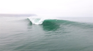 A medium-sized ocean wave curls and breaks in calm, open water under an overcast sky.