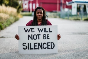 A person stands outdoors holding a sign that reads "WE WILL NOT BE SILENCED" in bold black letters.