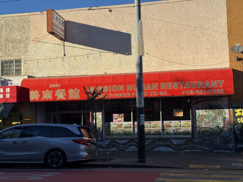 A restaurant with a red awning labeled "Mission Hunan Restaurant" and food photos displayed in its windows on a city street.