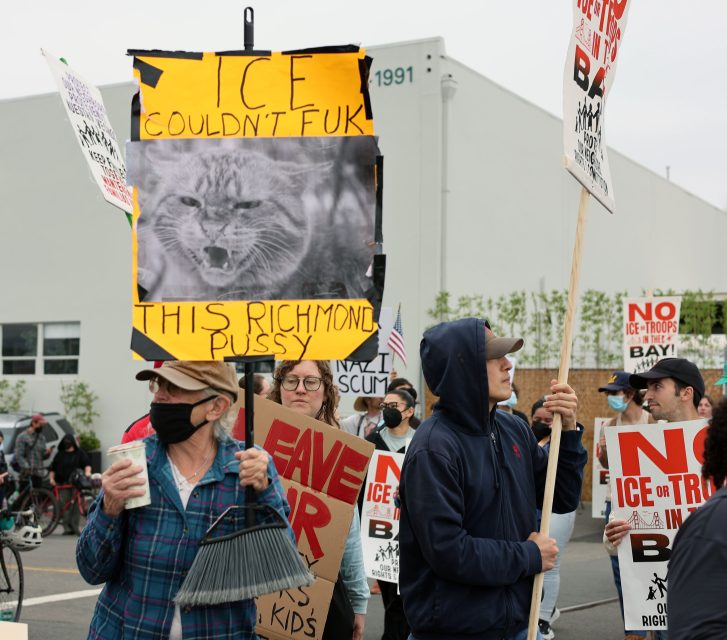 A group of protesters hold signs, including one with a cat image and text criticizing ICE, during a demonstration outside an industrial building.