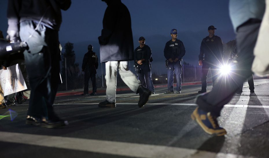 People walk past a line of police officers standing on a street at dusk, with one officer's flashlight pointed toward the camera.