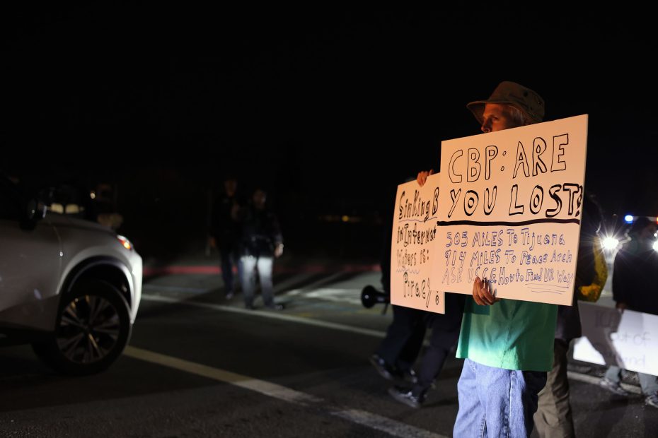 A person stands on a road at night holding a sign that reads "CBP: ARE YOU LOST?" while others gather nearby; a car is parked to the left.