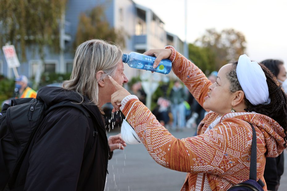 A woman in an orange patterned jacket pours water over another woman’s face on a street, likely to offer relief, with people and buildings visible in the background.