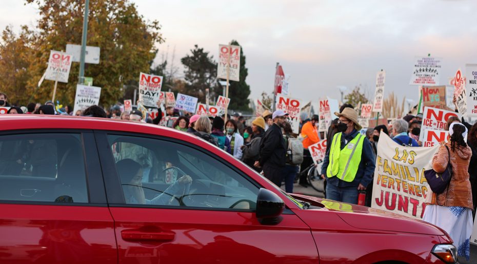 A red car passes by a large group of protesters holding signs with messages such as "NO ICE" and "NO BAY", while a few organizers in yellow vests stand nearby.