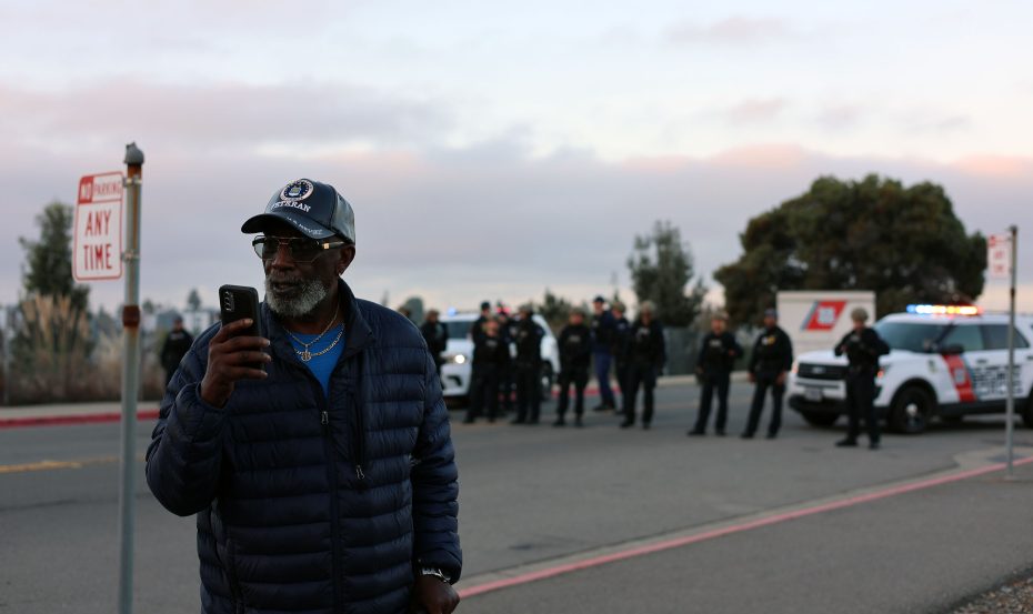 An older man in a blue jacket and cap uses his phone while police officers stand in a line near parked police cars in the background.