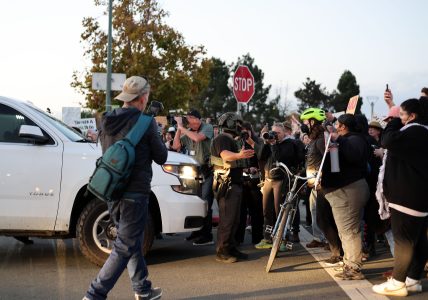 A group of people and police officers gather in front of a white SUV at a street intersection; several hold signs and cameras, and one person stands with a bicycle.
