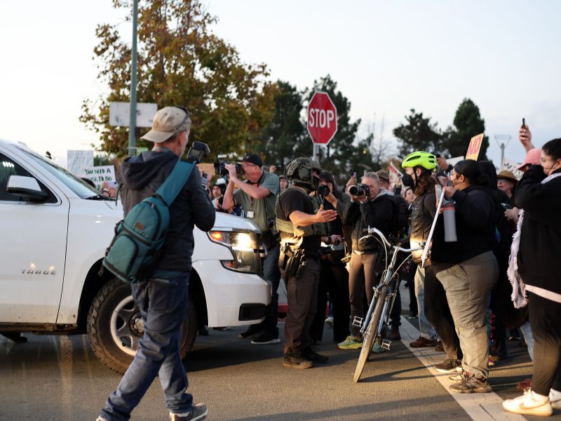 A group of people and police officers gather in front of a white SUV at a street intersection; several hold signs and cameras, and one person stands with a bicycle.
