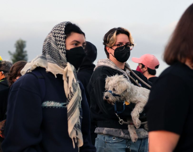 A group of people wearing face masks stand outdoors; one person holds a small white dog.