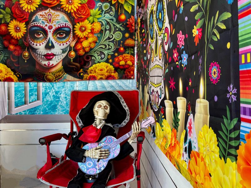 A skeleton figure holding a guitar sits in a red chair, surrounded by colorful Day of the Dead decorations featuring skull art, flowers, and candles.