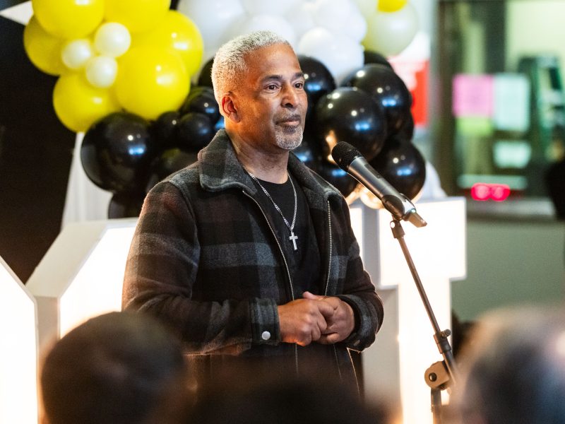 A man with short gray hair speaks into a microphone at an indoor event, standing in front of black, white, and yellow balloons.