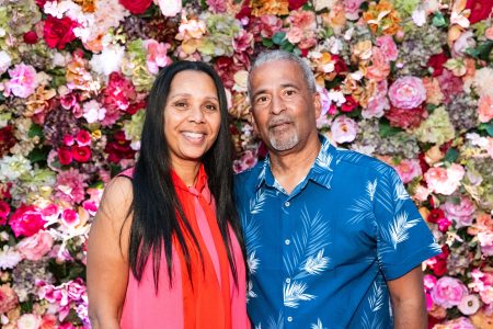 A woman and a man stand together in front of a colorful floral wall, both smiling at the camera.