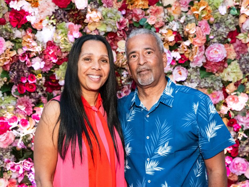 A woman and a man stand together in front of a colorful floral wall, both smiling at the camera.