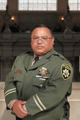 A man in a green San Francisco Sheriff's Department uniform stands indoors with his hands folded, facing the camera.