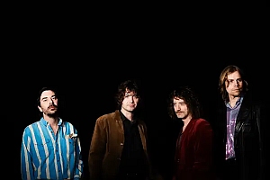 Four men stand in a row against a black background, each wearing different colored jackets and shirts, looking toward the camera.