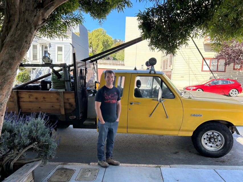 A person stands on the sidewalk next to a yellow Ford F-250 truck with wooden side panels and equipment in the bed, parked on a residential street.