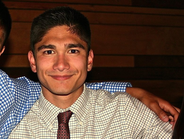 A man wearing a patterned dress shirt and red tie is smiling at the camera while someone’s arm rests on his shoulder.
