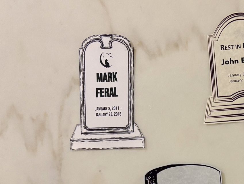 A paper illustration of a tombstone labeled "Mark Feral," showing dates January 8, 2011 – January 23, 2018, is attached to a marble surface.