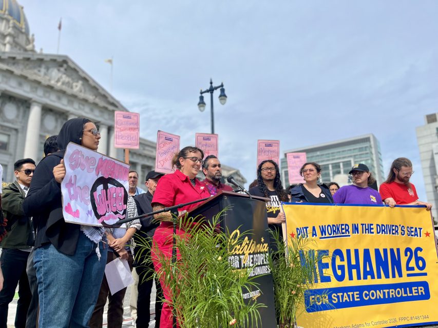A group of people stand at a podium during a rally; one holds a "Give Workers the Wheel" sign, another displays a "Meghann 26 for State Controller" banner.