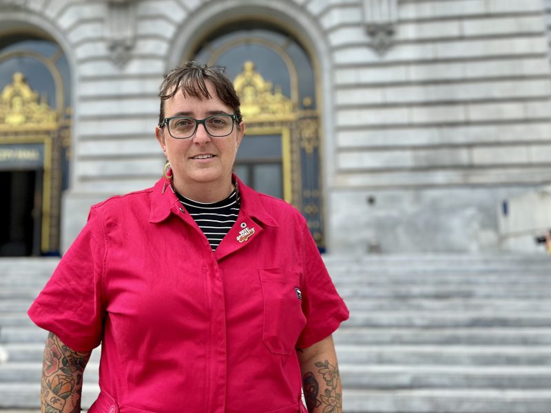 Person wearing glasses and a bright pink shirt stands in front of a building with ornate doors and stone steps.