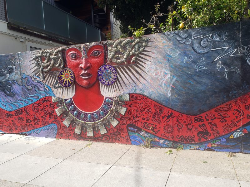 Mural of a red-skinned figure with ornate jewelry, large earrings, and patterned arms, set against a decorated background with abstract and nature motifs.