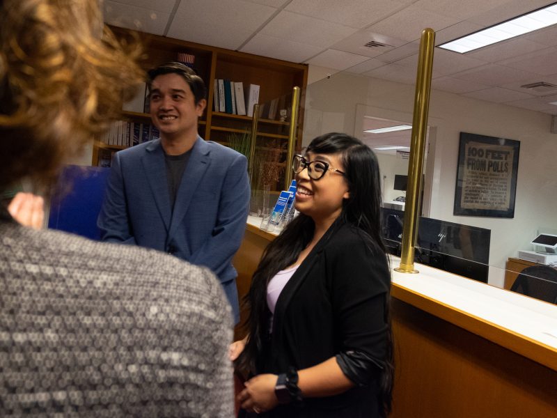 Two people standing and smiling in an office setting, talking to a person whose back is to the camera. A sign in the background reads "100 FEET FROM POLLS.
