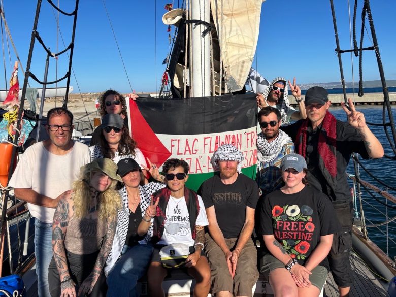A group of people pose on a boat, holding a large Palestinian flag with text. Some wear keffiyehs and themed shirts. The sea and a clear sky are visible in the background.